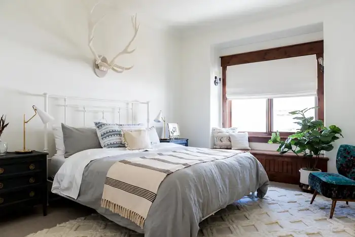 White Roman shade over a bedroom window beside a bed and green plant.
