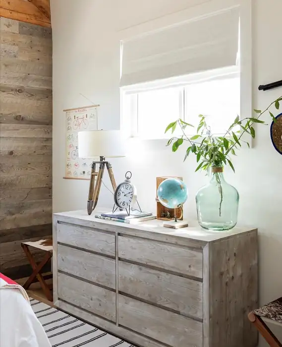 White Roman shade above a rustic dresser with a lamp and plants.